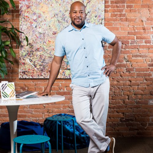 Man stands in office and rests his right hand on table next to him