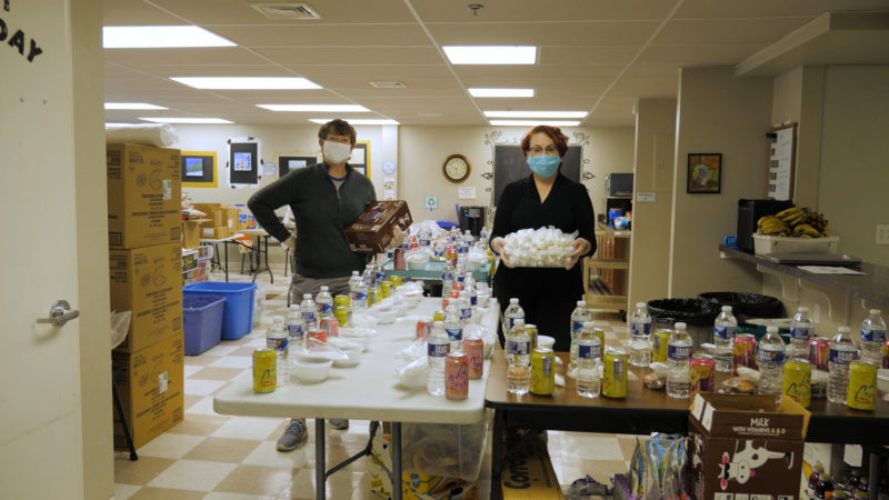 Two masked individuals stand behind tables with water and snacks