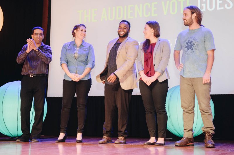 Group of people standing on a stage presenting. A projector screen is in the background