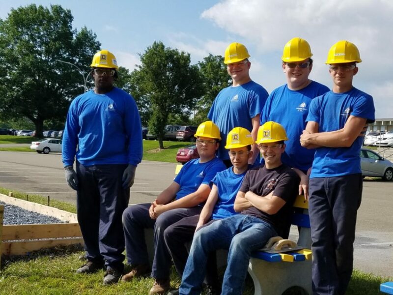 A group of people in hard hats stand and sit outside on a sunny day, looking at the camera
