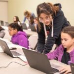 This picture shows girls working at computers, with a teacher standing near to help them. She is wearing a grey hoodie and the younger girls are wearing purple hoodies.