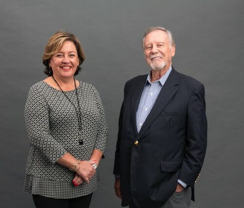 This picture shows two Caucasian people looking at the camera and smiling. The woman is wearing a grey shirt and long necklace. The man is wearing a blue sports jacket and light blue shirt.