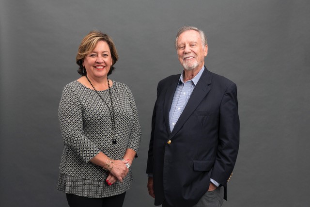 This picture shows two Caucasian people looking at the camera and smiling. The woman is wearing a grey shirt and long necklace. The man is wearing a blue sports jacket and light blue shirt.