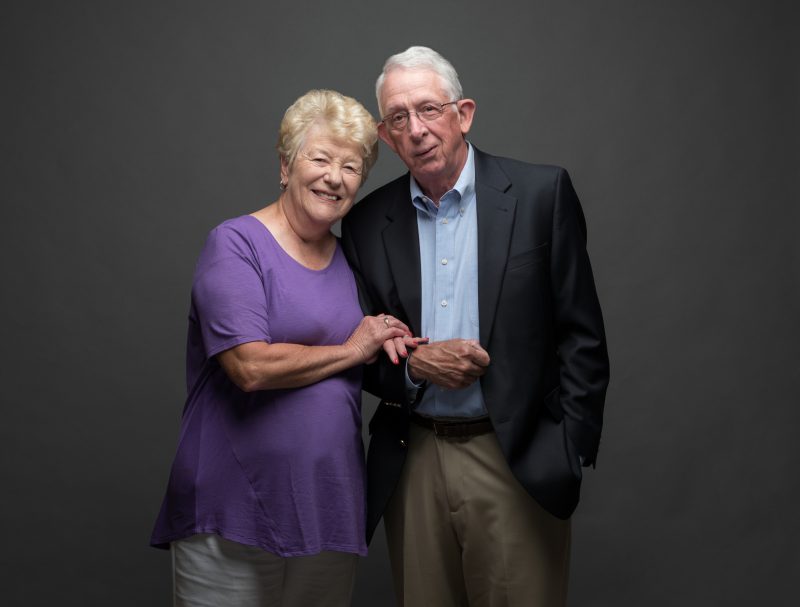 Married couple Sharon and Tom Kennedy stand close together, in front of a grey backdrop.