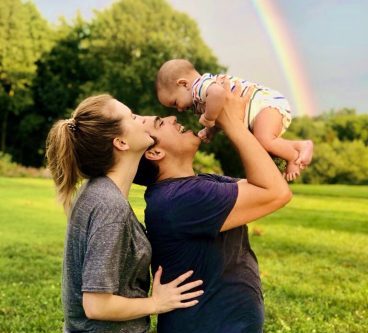 A family outside in a field. The husband is holding the baby and the wife is looking at the baby. There is a rainbow in the background.