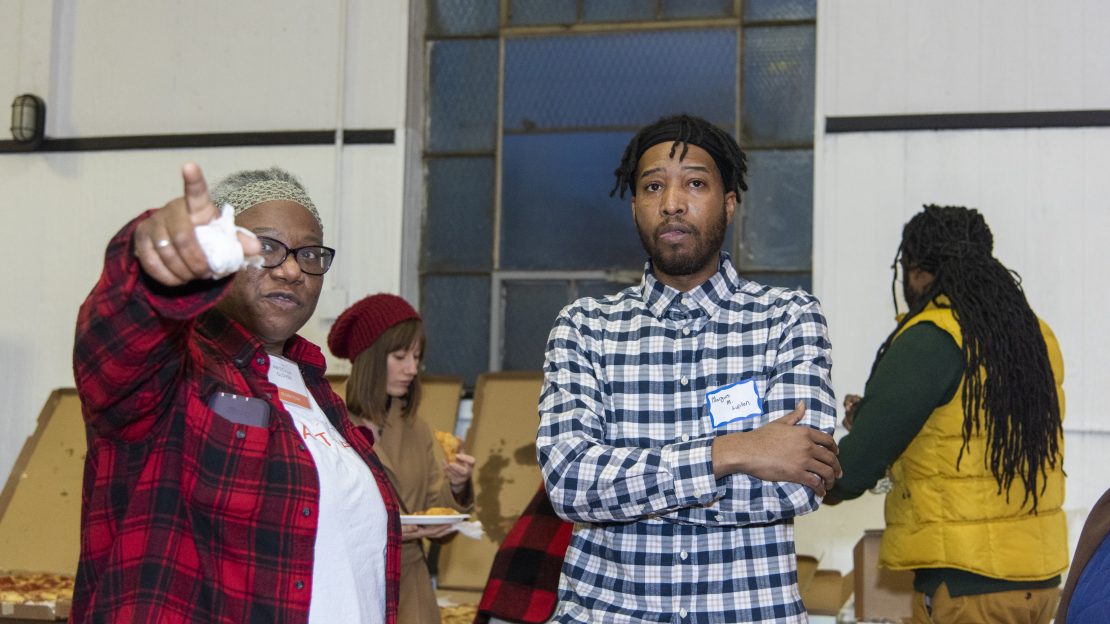 Two people stand next to each other while one points behind the camera