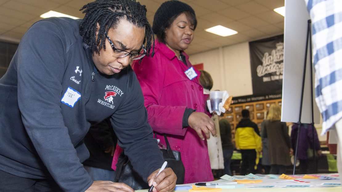 Two individuals stand in front of a table, writing on post its