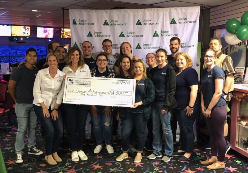 A group of people stand in front of a Junior Achievement sign, holding a large check.