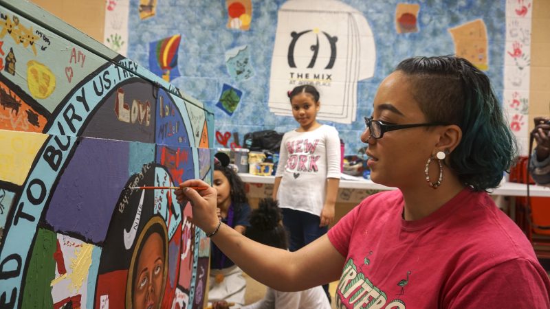 Woman painting on piano while another girl stands in the background and watches her.