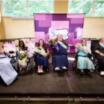 A group of women wearing fancy clothing and holding flowers. They are sitting in wheelchairs and smiling in front of a sign that says Ms. Wheelchair Pennsylvania.