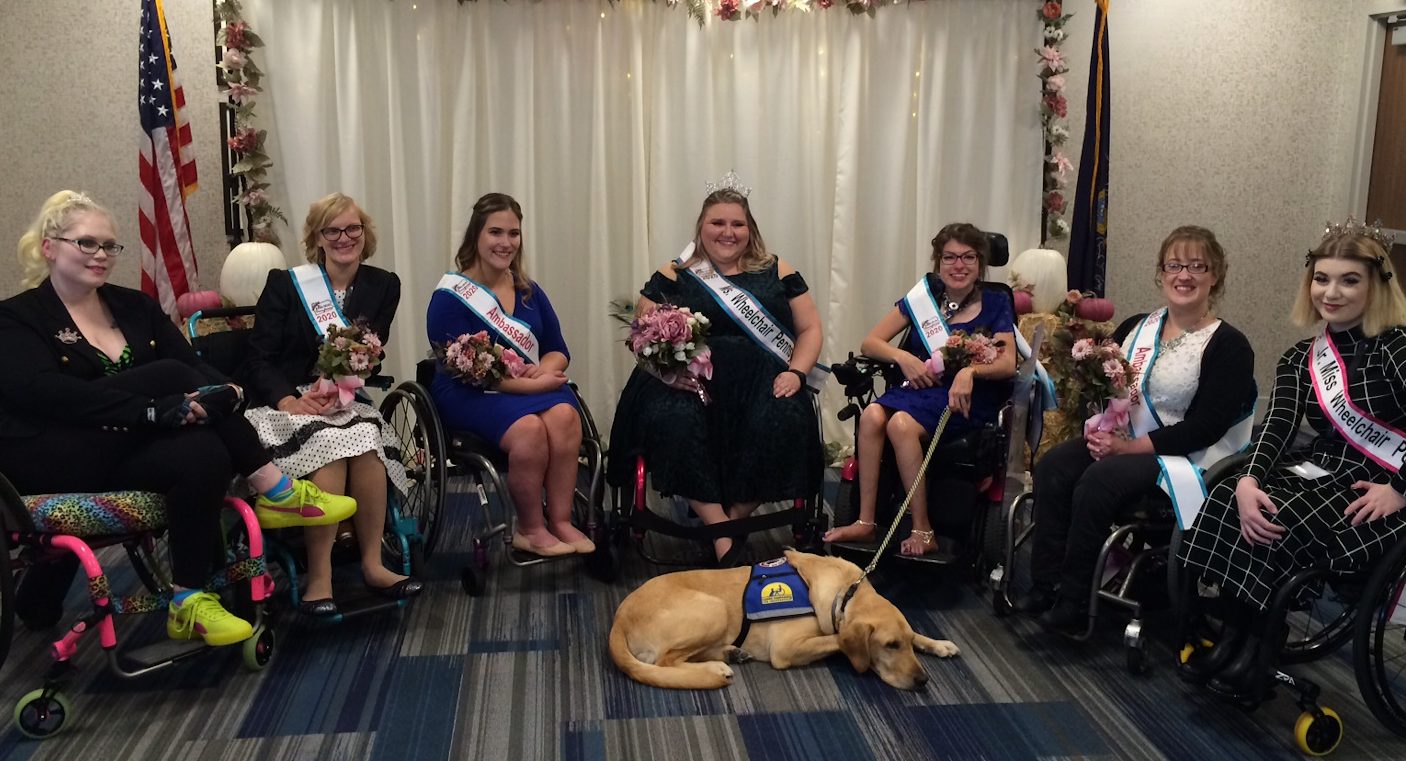 A group of women who use wheelchairs and a service dog face the camera. The women are wearing sashes that say "Ambassador" or "Ms. Wheelchair PA"