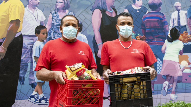 Two masked individuals stand in front of a mural holding crates of food