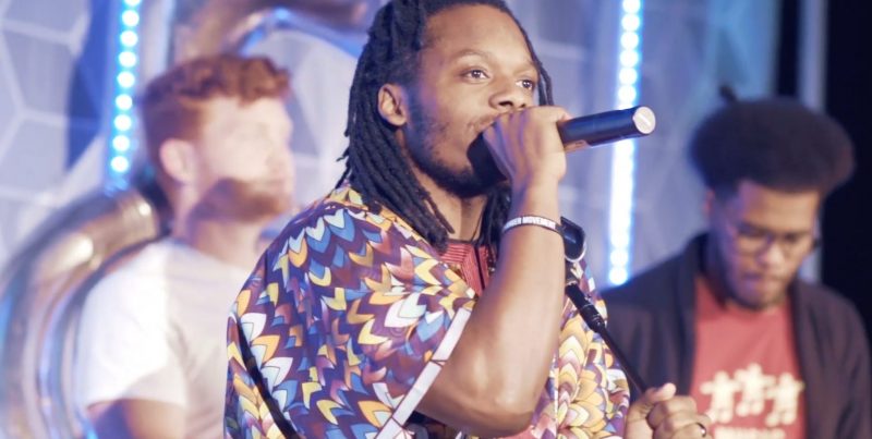 Photo of man holding a microphone. Two individuals are standing in the background.