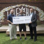 Three men stand in front of a brick wall outside, holding a large check that is a donation to Wildheart Ministries.