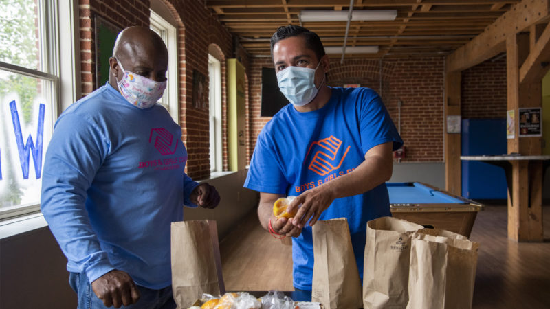 Two masked individuals are placing food into brown paper bags