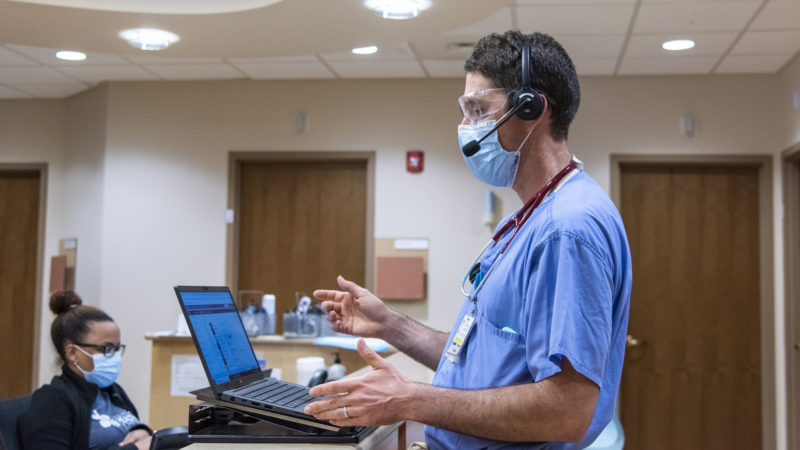 Masked doctor standing while looking at a computer. A woman is sitting in the bottom left corner.