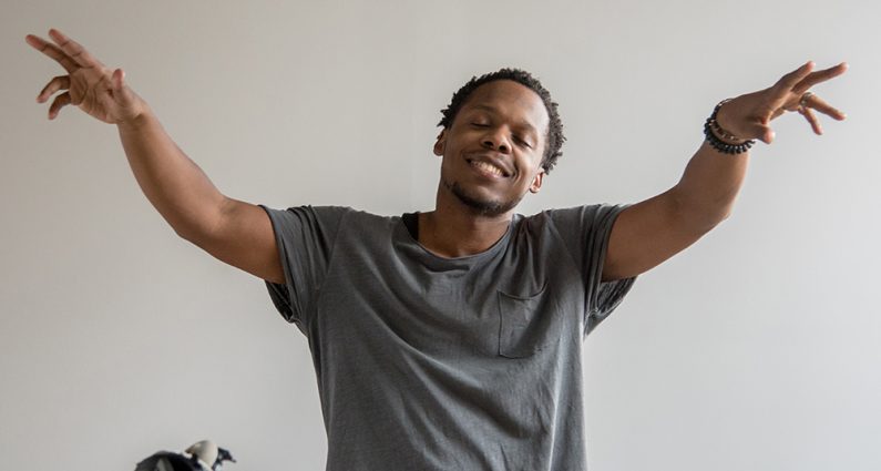 Portrait of Milan with his arms raised. Music and DJ equipment on table in front of him.
